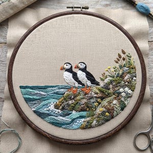May include: A circular embroidery hoop containing a needlepoint design of two puffins standing on a rocky cliffside overlooking a teal ocean with white-capped waves. The scene includes green vegetation, small yellow flowers, and white daisies.