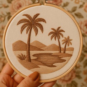Op de afbeelding: Geborduurd kunstwerk in een houten hoepel. Het ontwerp toont een strandscène met drie palmbomen, glooiende heuvels en een waterpartij, allemaal in bruintinten. De achtergrond is een neutrale beige stof.
