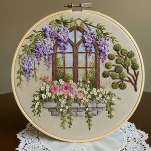 以下が含まれることがあります： 木製のフレームに入った刺繍アート。茶色の窓、紫色の藤、ピンクと白の花、緑の葉がデザインされています。背景はニュートラルなベージュ色の生地です。