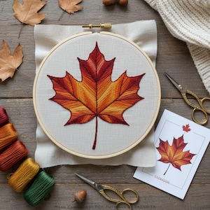 Könnte beinhalten: Ein Stickrahmen mit einem bunten Herbstblatt-Design in Rot-, Orange- und Gelbtönen. Das Blatt ist auf cremefarbenem Stoff gestickt, vor einem Holzuntergrund. Zusätzliche Elemente sind Stickgarn, Scheren und eine Karte mit einer passenden Blattillustration.