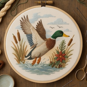 Puede incluir: Un bordado detallado en un aro de madera que representa un pato real en vuelo sobre el agua. El pato tiene plumas marrones, verdes y blancas, con un pico amarillo y patas naranjas. La escena incluye juncos, una flor roja y un cielo azul con nubes.