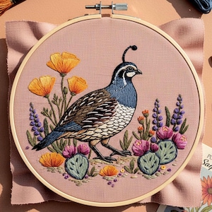 Op de afbeelding: Geborduurde hoepelkunst met een kwartel met blauwe, bruine en witte veren, omringd door oranje klaprozen, paarse wilde bloemen en groene cactussen. Het kunstwerk is geplaatst op een roze stoffen achtergrond, omlijst in een houten hoepel.