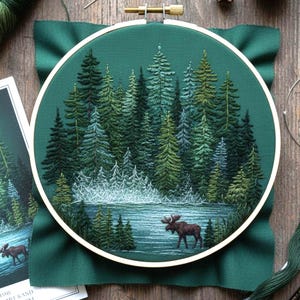 May include: An embroidered forest scene with evergreen trees, a lake, and a moose, set within a white embroidery hoop. The artwork is stitched on dark green fabric. The colour palette includes shades of green, blue, and brown, creating a detailed landscape.