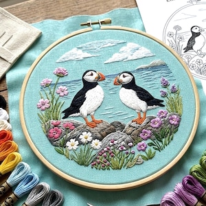 May include: A completed embroidery hoop featuring two puffins standing on rocks, with a blue ocean and sky background. The embroidery includes pink, purple, and white flowers, and green foliage. The hoop is made of wood.
