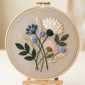 May include: A circular wooden embroidery hoop containing a beige fabric panel with a hand-stitched floral design. The embroidery features white, pink, and blue flowers, green leaves, and tan wheat stalks arranged in a central bouquet pattern.