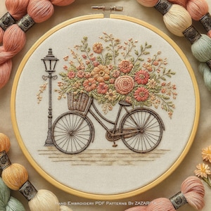 Puede incluir: Bordado en un aro de madera que representa una bicicleta con una cesta llena de flores y una farola. El diseño utiliza hilos en tonos melocotón, naranja y verde. Ovillos de hilo de colores similares rodean la obra.