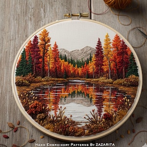 May include: Hand-stitched embroidery art in a wooden hoop. The artwork depicts a serene autumn landscape with a lake reflecting colorful trees in shades of red, orange, and green. Mountains are visible in the background. The text "Hand Embroidery Patterns By Zazarita" is at the bottom.