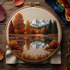 May include: A hand-stitched landscape scene in a wooden embroidery hoop. The design depicts a lake reflecting autumn trees and snow-covered mountains. The embroidery uses shades of orange, red, green, and blue. The text at the bottom reads "Hand Embroidery PDF Patterns By Zazarita."