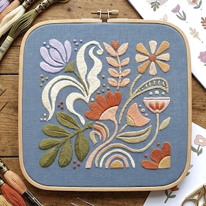 May include: A square embroidery hoop with a blue fabric featuring a floral design. The embroidery includes flowers, leaves, and a rainbow in shades of orange, peach, green, and white. The wooden hoop is visible.