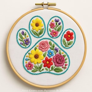 May include: A hand-embroidered floral paw print design on white fabric, set within a wooden embroidery hoop. The paw print is filled with colorful flowers, including yellow sunflowers, red and pink roses, and blue accents. The text "Hand Embroidery Patterns By Zazarita" is at the bottom.