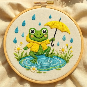 May include: Embroidered artwork in a wooden hoop features a green frog wearing a yellow raincoat and holding a yellow umbrella. The frog stands in a blue puddle with embroidered raindrops and flowers. The background is a cream-colored fabric.