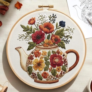 May include: Embroidered floral teapot design in a wooden hoop. The teapot is adorned with colorful flowers in shades of red, orange, yellow, and green. The background is a neutral white fabric.