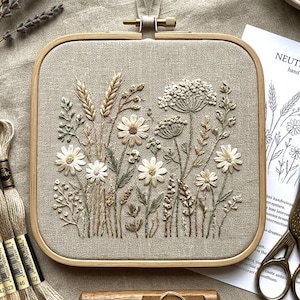 May include: A square embroidery hoop with a beige linen fabric featuring a detailed floral design. The embroidery showcases various wildflowers in shades of white, beige, and green, with a wooden frame and hanging hardware.