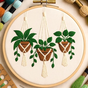 May include: Embroidered hoop art featuring three hanging planters with green leaves and brown pots. The pots are suspended by cream-colored macrame cords and tassels. The embroidery is set against a cream-colored fabric background, framed by a wooden hoop.