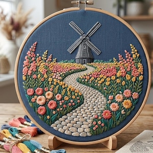 May include: Embroidered artwork in a wooden hoop. The design features a windmill, a cobblestone path, and a field of colorful flowers in shades of pink, yellow, and orange against a dark blue background. Embroidery floss in various colors is visible.
