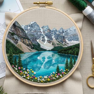 Può includere: Arte paesaggistico ricamato in un telaio di legno. La scena raffigura una catena montuosa con cime innevate che si riflettono in un lago turchese. Il primo piano presenta fiori selvatici colorati e alberi sempreverdi. Il ricamo è dettagliato e vibrante.