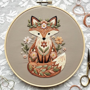 May include: Embroidered fox art in a wooden hoop. The fox is depicted with brown, orange, and cream-colored thread, adorned with floral details and a leafy crown. The background is a neutral tan fabric, and the hoop is approximately 8 inches in diameter.