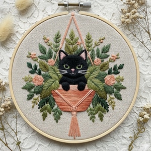 Może przedstawiać: Haft na tamborku przedstawiający czarnego kota w makramowym kwietniku otoczonym zielonymi liśćmi i brzoskwiniowymi różami. Kot ma zielone oczy i białe wąsy. Tamborek jest drewniany.
