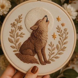 May include: An embroidered artwork in a wooden hoop depicts a brown wolf howling at a textured, cream-coloured moon. The design includes brown foliage, yellow stars, and a cream-coloured background. The artwork is a nature-themed piece.