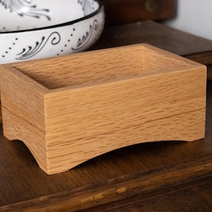 Puede incluir: Una caja rectangular de madera clara, con base curvada. Está sobre una superficie de madera oscura. En el fondo, un cuenco decorativo con un diseño en blanco y negro.