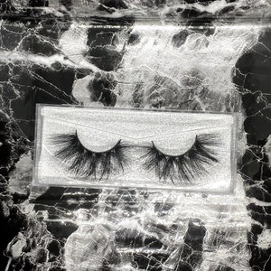 May include: A pair of black, fluffy false eyelashes in a clear plastic case. The lashes are densely packed and have a dramatic, voluminous appearance. The case sits on a black and white marble-patterned surface.