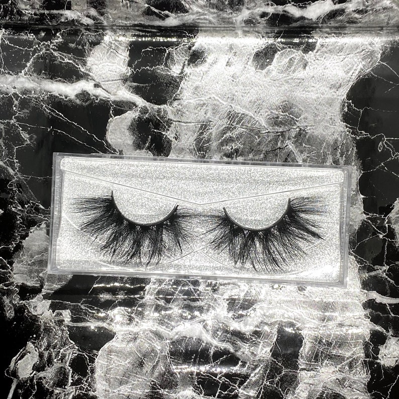 May include: A pair of black, fluffy false eyelashes in a clear plastic case. The lashes are densely packed and have a dramatic, voluminous appearance. The case sits on a black and white marble-patterned surface.