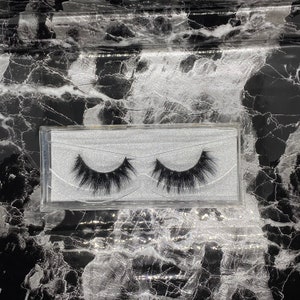 May include: A pair of black false eyelashes in a clear plastic case. The lashes are thick and full, with a natural-looking curl. The case sits on a black and white marble patterned surface.