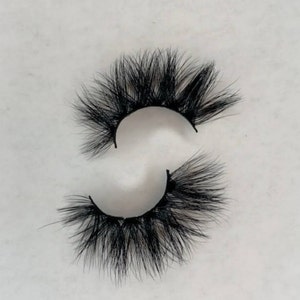 May include: A pair of dramatic, black false eyelashes. The lashes are thick and full, with a layered design that creates a voluminous effect. The individual hairs are long and tapered, giving a wispy appearance. The lashes are designed to be applied to the upper eyelid.