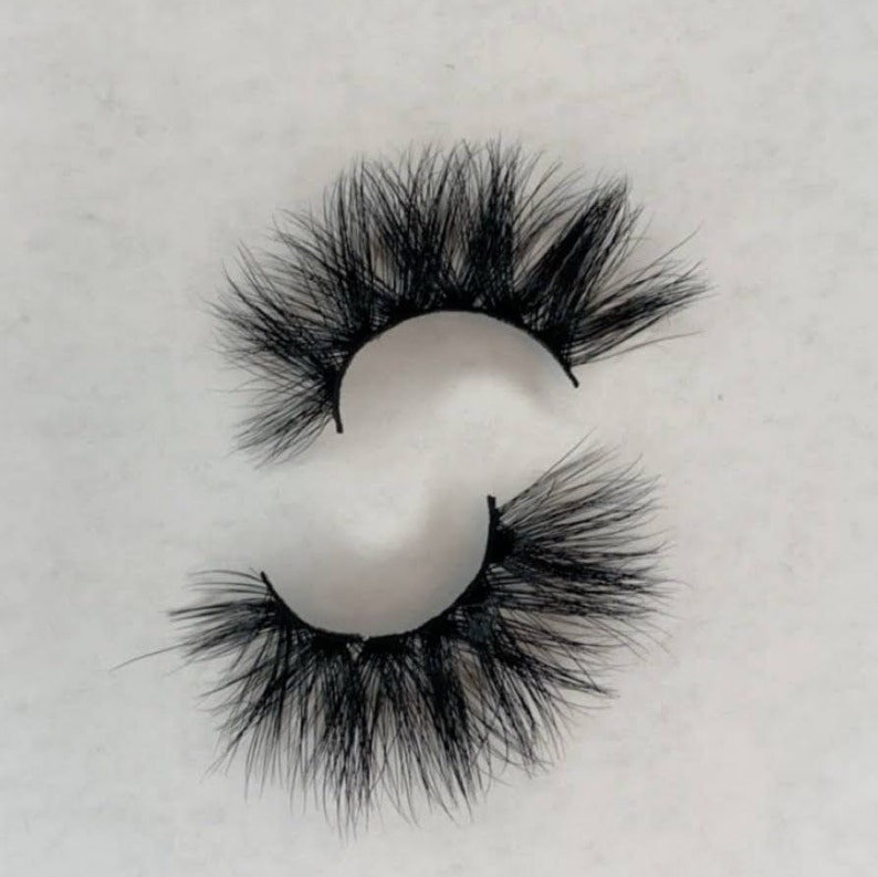 May include: A pair of dramatic, black false eyelashes. The lashes are thick and full, with a layered design that creates a voluminous effect. The individual hairs are long and tapered, giving a wispy appearance. The lashes are designed to be applied to the upper eyelid.