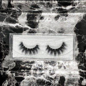 May include: A pair of black false eyelashes in a clear plastic case. The lashes are long and fluffy, with a natural-looking curl. The case sits on a black and white marble-patterned surface.