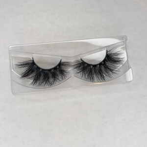 May include: A pair of black, fluffy false eyelashes in a clear plastic case. The lashes are thick and voluminous, with a curved shape. The case is rectangular with a molded design to hold the lashes.