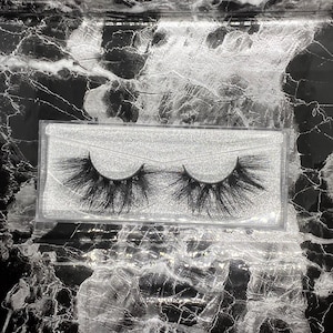 May include: A pair of black false eyelashes in a clear plastic case. The lashes are long and full, with a dramatic, layered design. The case sits on a black and white marble-patterned surface.