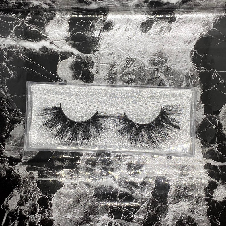 May include: A pair of black false eyelashes in a clear plastic case. The lashes are long and full, with a dramatic, layered design. The case sits on a black and white marble-patterned surface.