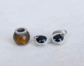 Könnte beinhalten: Eine facettierte, bernsteinfarbene Perle mit silberfarbenem Sockel liegt neben einer silberfarbenen Sonnenbrillenbrosche. Die Sonnenbrille hat schwarze Gläser und ist mit kleinen, klaren Kristallen besetzt. Die Brosche hat ein strukturiertes Design.