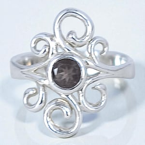 May include: A silver ring with a dark gemstone center. The ring features an ornate, swirling design around the gemstone. The band is smooth and simple, contrasting with the detailed top. The gemstone is round and set in a bezel.