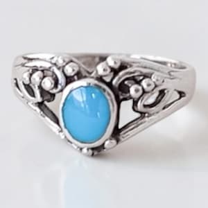 May include: A silver ring featuring a central oval, light blue gemstone. The ring showcases an intricate, openwork design with floral and beaded embellishments. The band is smooth and polished, set against a white backdrop.