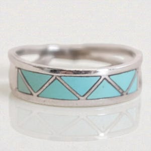 Puede incluir: Un anillo plateado con una banda que presenta un diseño geométrico de triángulos turquesa. Los triángulos están delineados en un color más oscuro, creando un contraste con la banda plateada. El anillo se muestra sobre una superficie reflectante.