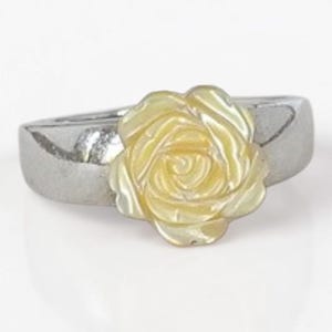 May include: A silver-toned metal ring featuring a central, carved, pale yellow rose ornament. The rose has a smooth, iridescent finish with layered petals, while the band is a solid, polished silver-coloured metal with a smooth, rounded surface.