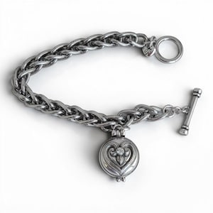 May include: A silver-toned metal bracelet featuring a thick, braided chain design. The bracelet includes a toggle clasp and a round, hinged locket charm decorated with an embossed heart pattern and a single small, clear embedded crystal.
