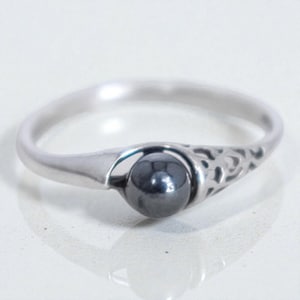 May include: A silver ring with a dark gray, spherical stone. The ring has a simple band with a decorative openwork design on one side of the stone setting. The stone is approximately 0.25 inches in diameter.