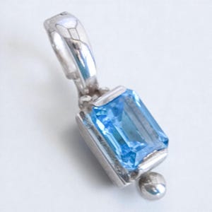 May include: A silver pendant with a rectangular, light blue gemstone. The pendant has a small, round accent at the bottom and a loop at the top for a chain. The gemstone is set in a silver frame.