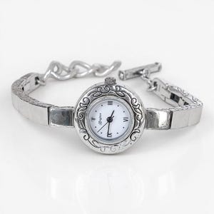 May include: A silver-toned Brighton watch with a white face and black hands, featuring Roman numerals and a decorative bezel. The watch has a matching silver bracelet with a toggle clasp.