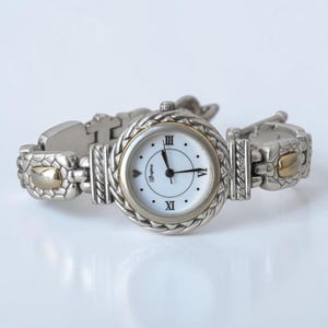 May include: A silver-tone analog wristwatch with a white face, black hands, and Roman numerals at the three, six, and nine o'clock positions. The bezel and bracelet feature a braided metal texture with gold-tone accents on the links.