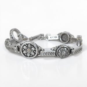 May include: A silver-toned metal link bracelet featuring alternating circular medallions with embossed floral and textured patterns, connected by rectangular segments with etched leaf motifs, finished with a toggle clasp closure.