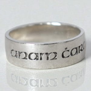 May include: A silver ring with the inscription "anam cara" engraved around the band. The ring has a smooth, polished finish and a simple, classic design. The ring is likely made of sterling silver.