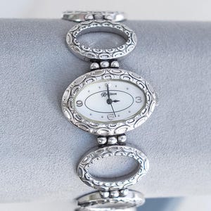 Könnte beinhalten: Eine silberfarbene Analoguhr mit ovalem Zifferblatt in Weiß, schwarzen Zeigern und dem Schriftzug Brighton. Das Armband besteht aus miteinander verbundenen ovalen Gliedern mit einem strukturierten Prägemuster und kleinen Silberkugeln.