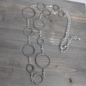 Può includere: Collana argentata con una serie di cerchi aperti interconnessi di diverse dimensioni. I cerchi sono collegati da piccole catene e presentano un motivo strutturato sui bordi. La collana è esposta su una superficie di legno grigio.