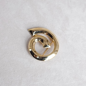 May include: A gold-toned brooch in the shape of a spiral. The spiral is made of a single piece of metal and has a smooth, polished finish.