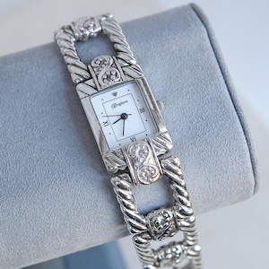 May include: A silver-toned Brighton watch with a rectangular white face and Roman numeral hour markers. The watch features a decorative link bracelet and is displayed on a gray surface. The watch face has the Brighton logo.
