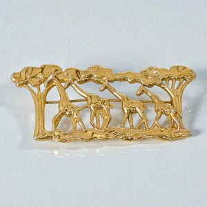 Könnte beinhalten: Goldfarbene Brosche mit einer stilisierten Szene von Giraffen und Bäumen. Die Brosche zeigt vier Giraffen unter Bäumen, alle in einem detaillierten, dreidimensionalen Design. Die Brosche hat eine rechteckige Form.